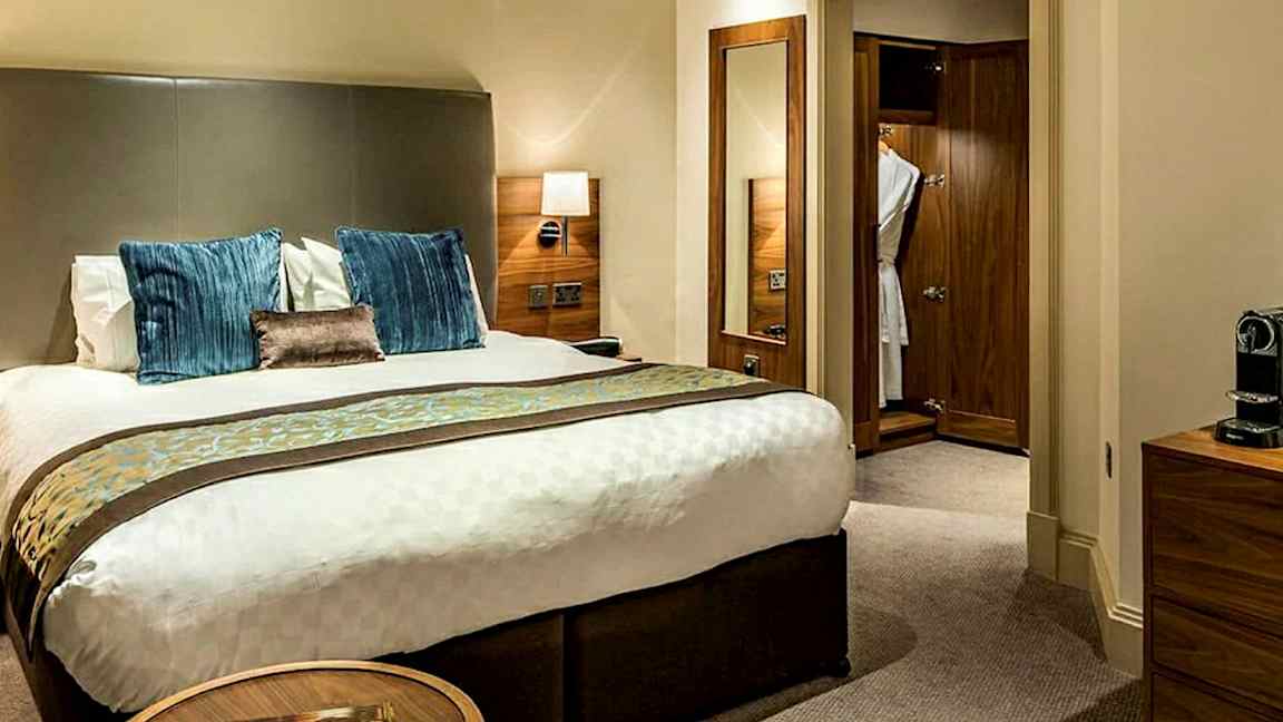 Kuva hotellihuoneesta, jossa on parivuode, yöpöytä ja puinen vaatekaappi. (Tekoälyn luoma teksti)