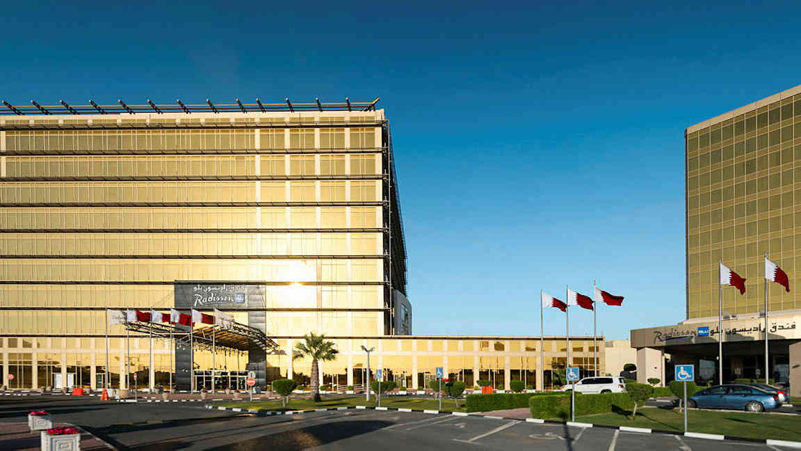 Moderni rakennus, jossa on kultaväriset lasiseinät, lipputankoja ja pysäköintialue. (Tekoälyn luoma teksti)