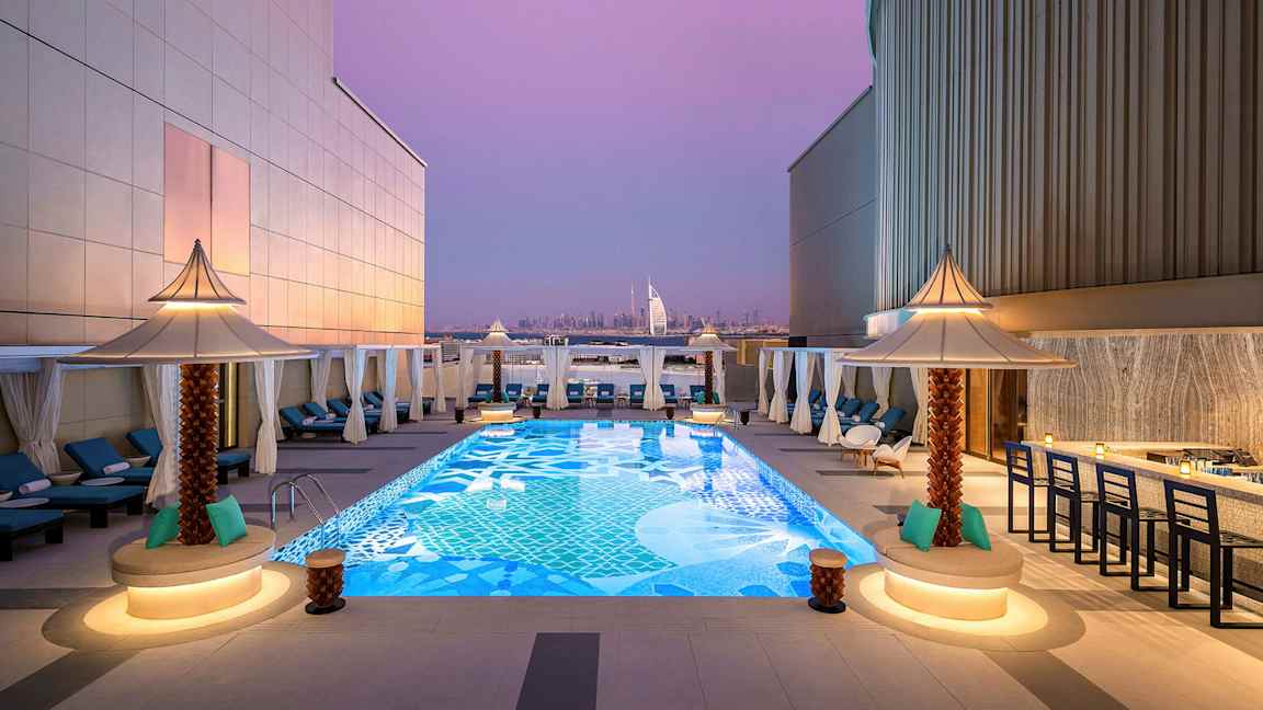 Kattoterassi, jossa koristeellinen uima-allas, jota ympäröivät aurinkotuolit ja pienet aurinkovarjot. Taustalla näkyy Burj Al Arab ja Dubain kaupunkimaisema violetin iltataivaan alla. (Tekoälyn luoma teksti)
