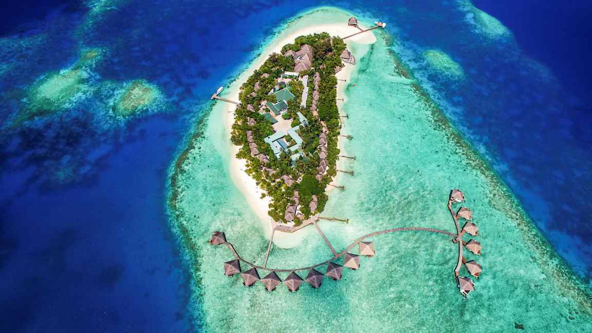 Ilmakuva trooppisesta saaresta, jossa rakennuksia turkoosinsinisen veden ympäröimänä. (Tekoälyn luoma teksti)