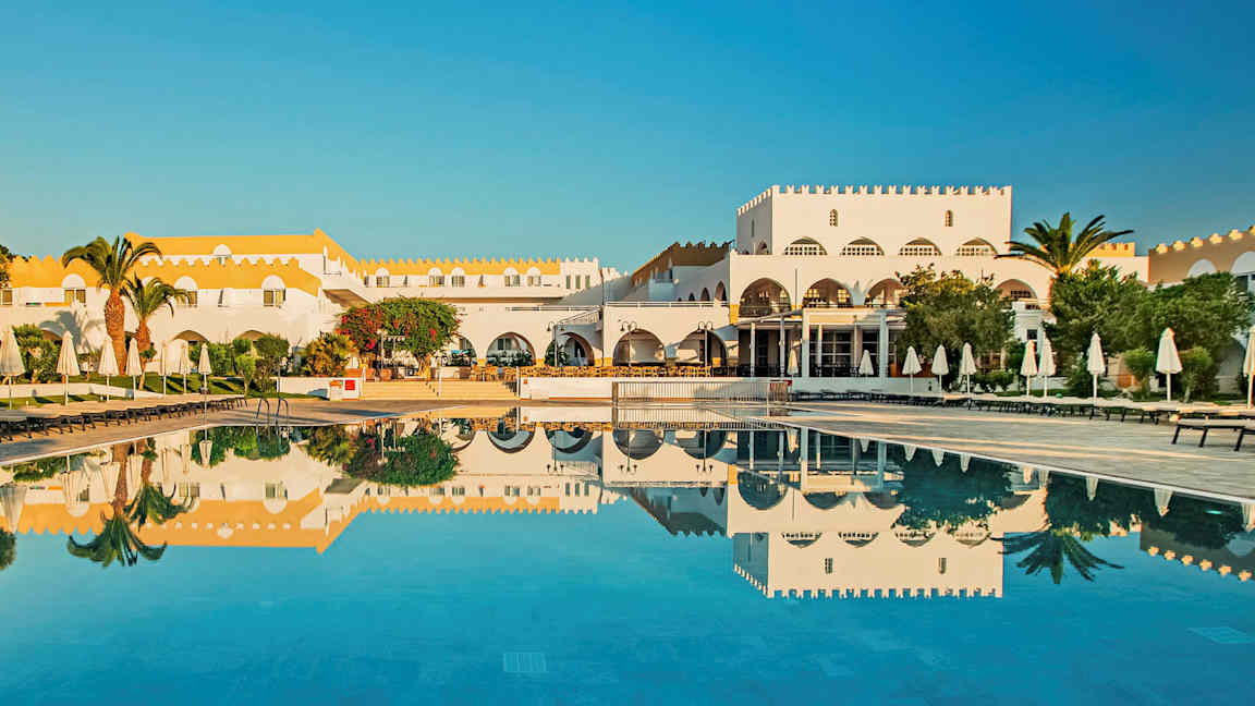 Valkoinen rakennus, jossa keltainen katto ja uima-allas, ympärillä aurinkotuoleja ja varjoja. (Tekoälyn luoma teksti)