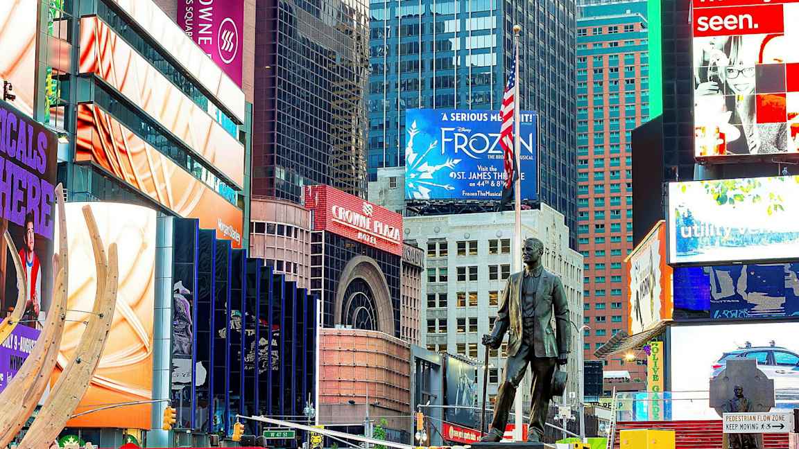 Kaupungin aukio, jossa värikkäitä kyltejä, mainoksia ja patsas etualalla. (Tekoälyn luoma teksti)