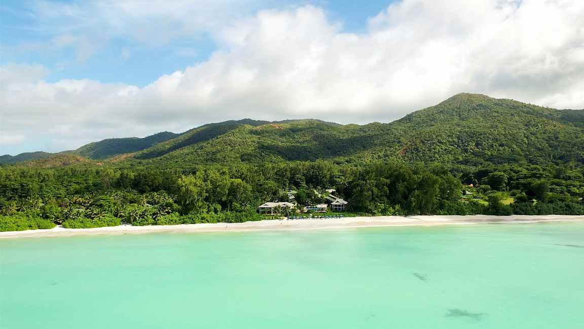 Ranta, jossa on valkoista hiekkaa, metsän peittämä vuorijono ja turkoosia vettä. (Tekoälyn luoma teksti)