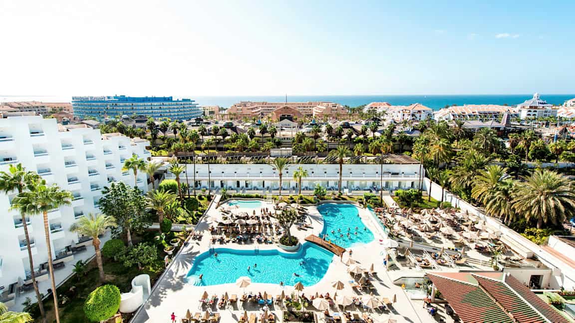 Näkymä hotellialueelle, jossa uima-altaita, aurinkotuoleja, palmuja ja rakennuksia taustalla. (Tekoälyn luoma teksti)