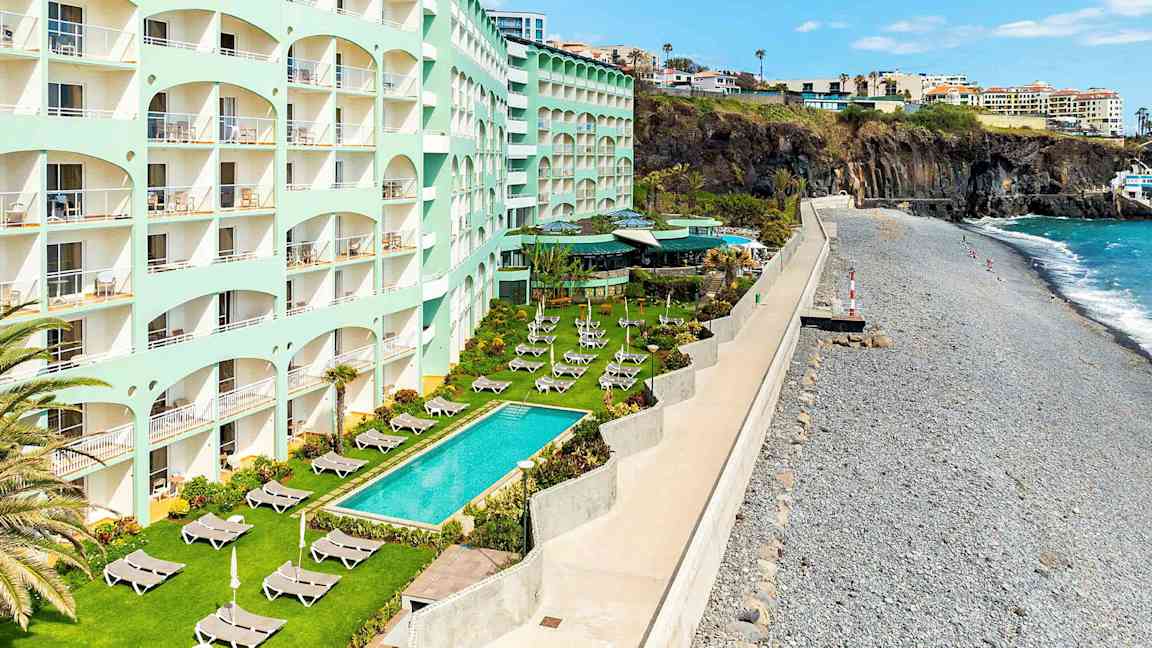 Hotelli, jossa on useita kerroksia meren äärellä, nurmikko aurinkotuoleilla ja uima-allas ulkona. (Tekoälyn luoma teksti)