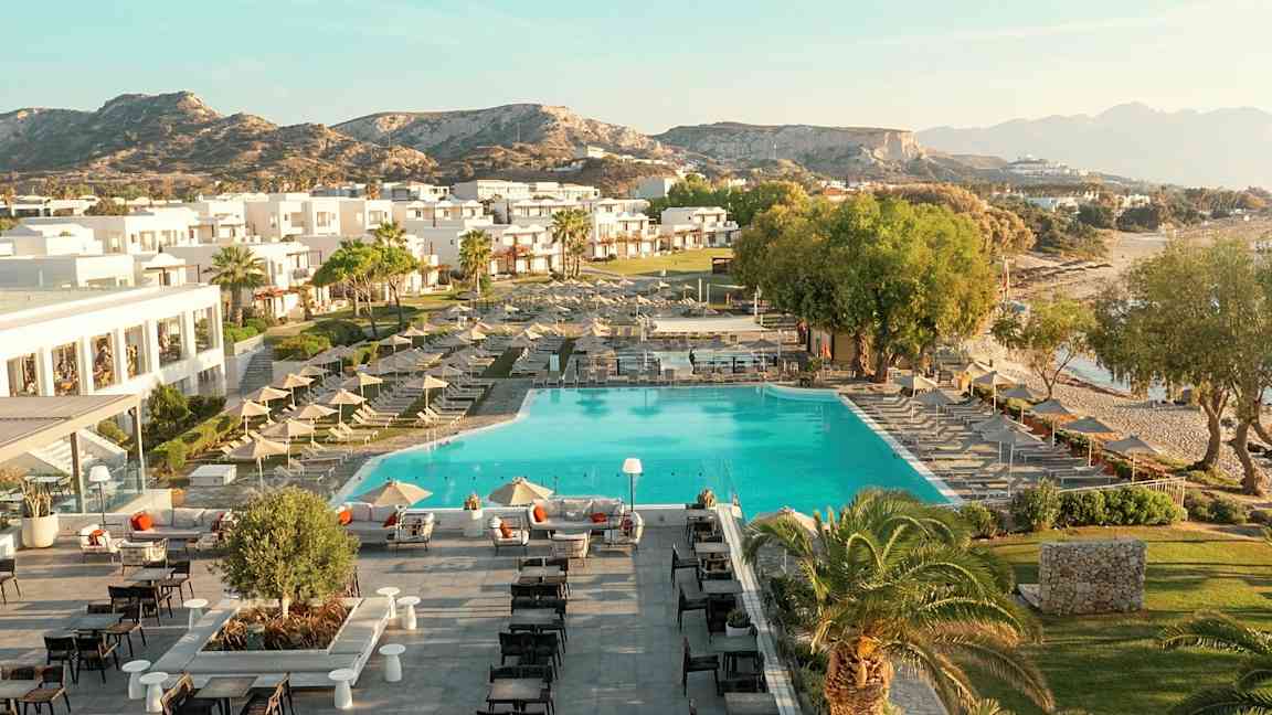 Ilmakuva lomakeskuksesta, jossa uima-allas, aurinkotuoleja, rakennuksia ja ympäröivä maisema. (Tekoälyn luoma teksti)