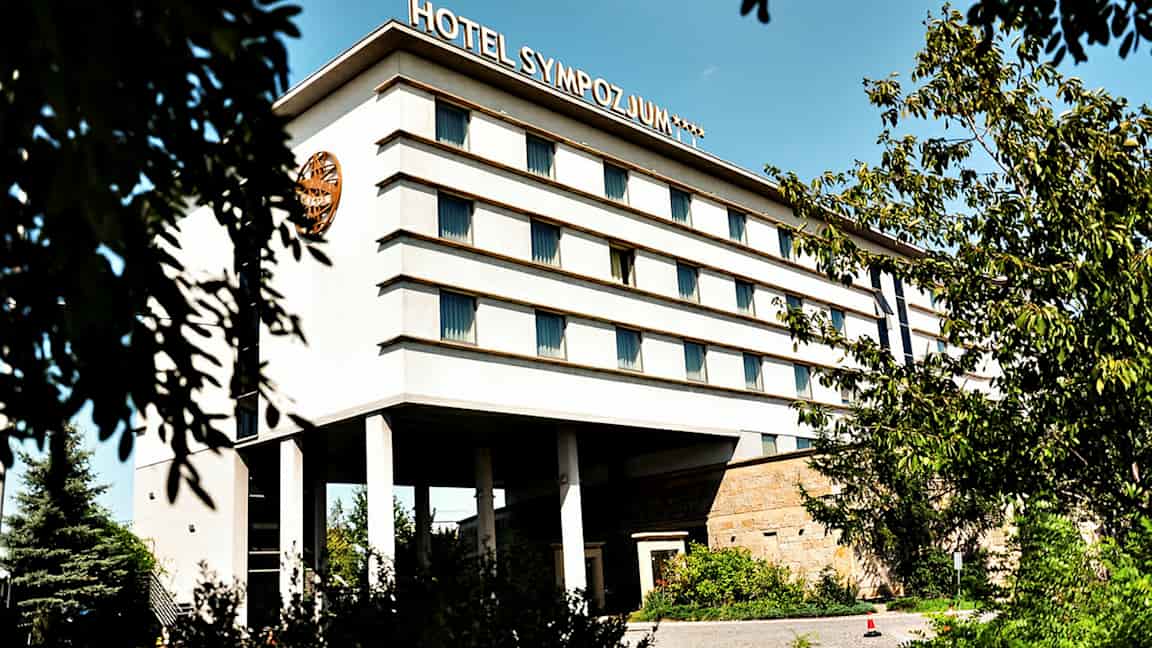 Monikerroksinen rakennus, jonka kyljessä lukee 'Hotel Sympozjum', ympäröitynä puilla ja kasvillisuudella. (Tekoälyn luoma teksti)