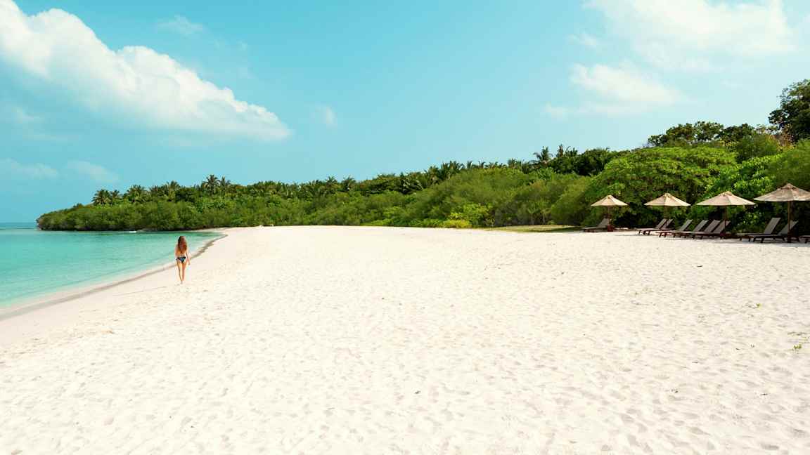 Nainen kävelee hiekkarannalla, jossa taustalla turkoosi vesi ja vihreä kasvillisuus. (Tekoälyn luoma teksti)