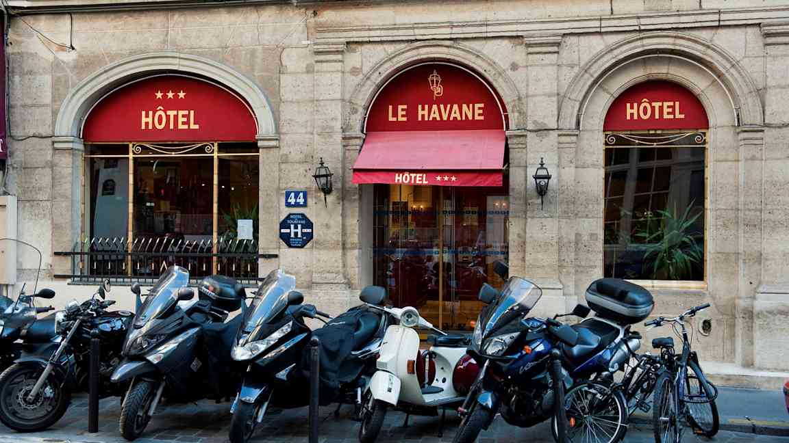 Kuva, joka esittää hotellin sisäänkäyntiä. Nimi Le Havane ja kolme tähteä näkyvät markiisissa. Rakennuksen edessä on pysäköityjä moottoripyöriä ja polkupyöriä. (Tekoälyn luoma teksti)