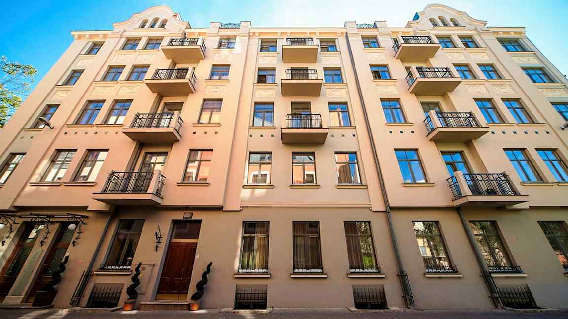 Beigen rakennuksen julkisivu, jossa on useita parvekkeita ja siniset ikkunat. (Tekoälyn luoma teksti)