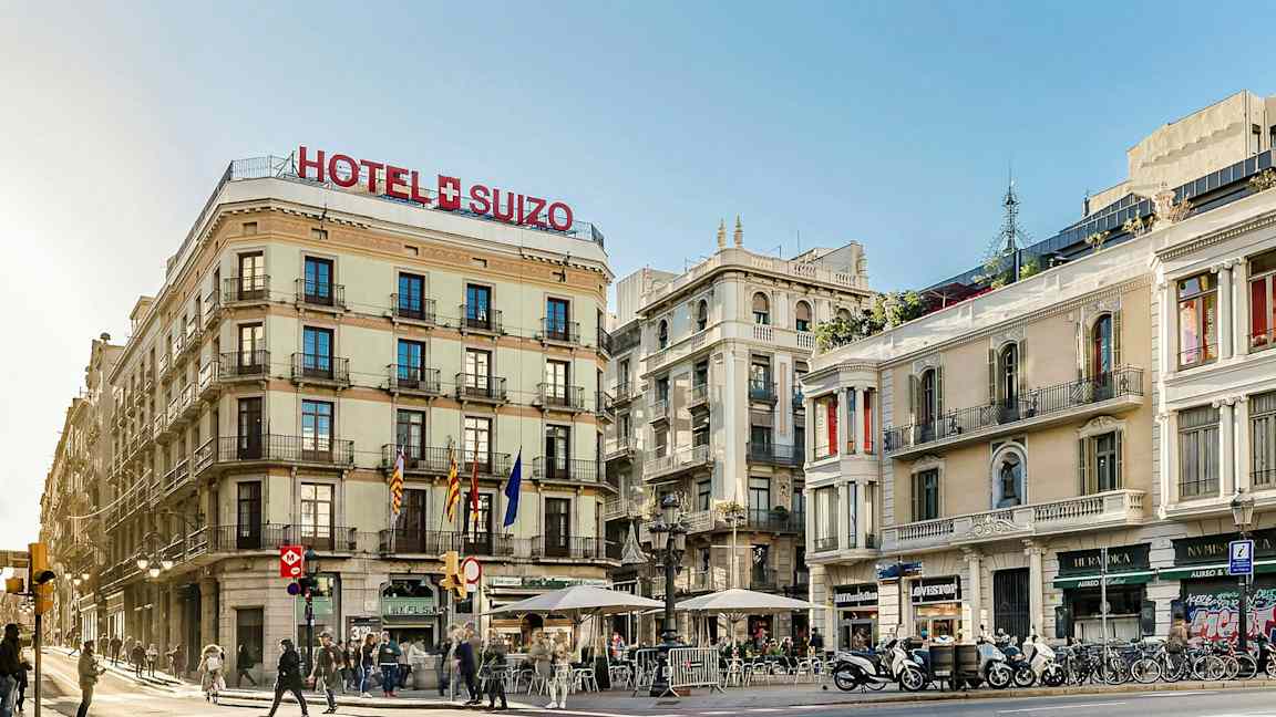 Kadunkulma, jossa on rakennuksia, joista yhdessä lukee hotelli Suizo. Muutamia ihmisiä kulkee ja istuu ulkoterasseilla. (Tekoälyn luoma teksti)