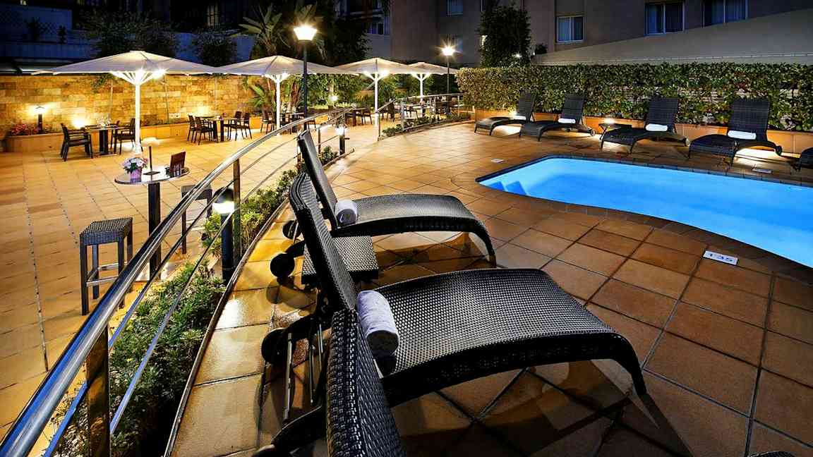 Hotellin allasalue, jossa aurinkotuoleja ja aurinkovarjoja valaistulla laatoitetulla ulkoalueella. (Tekoälyn luoma teksti)