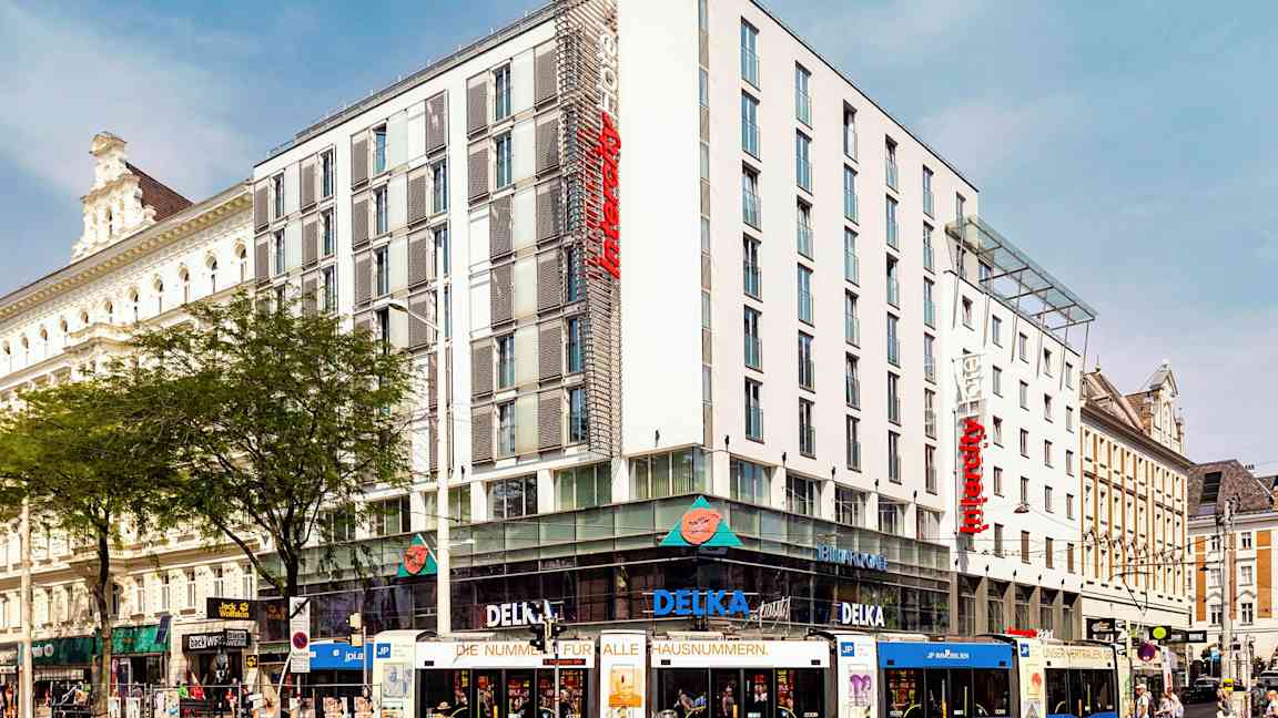 Monivärinen raitiovaunu modernin hotellin edessä kaupunkimaisemassa. (Tekoälyn luoma teksti)