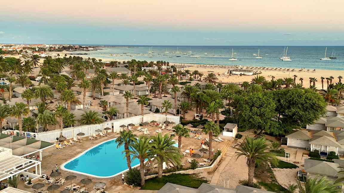 Näköala lomakeskukseen, jossa on uima-allas, palmuja ympärillä ja rannan lähellä, jossa veneitä meressä. (Tekoälyn luoma teksti)