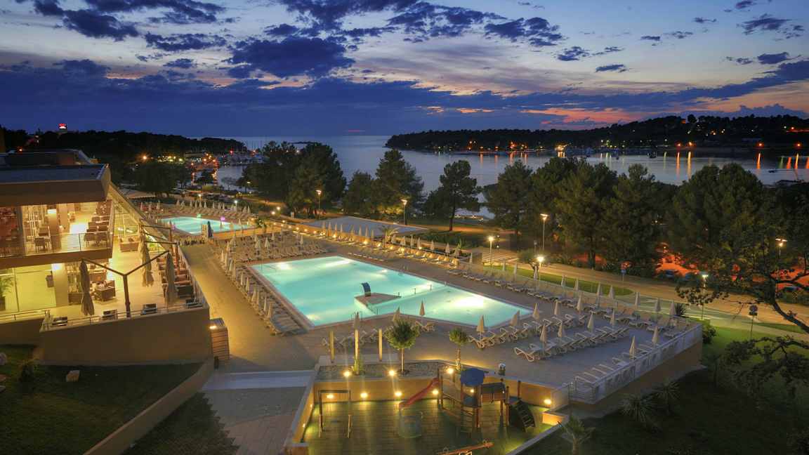 Iltanäkymä hotellialueesta valaistuine uima-altaineen, ympäröivine terasseineen ja puineen, taustalla on järvi. (Tekoälyn luoma teksti)