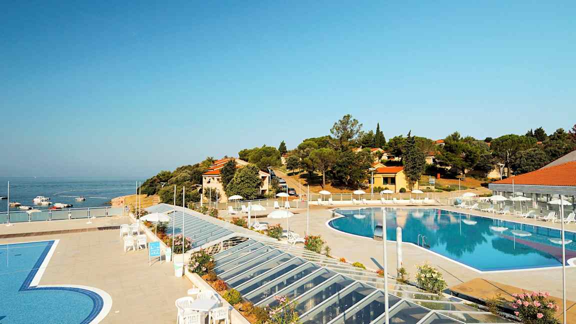 Ulkoallas, jota ympäröi terassi, jossa on aurinkotuoleja ja aurinkovarjoja. Taustalla näkyvät meri sekä rakennukset ja kasvillisuus. (Tekoälyn luoma teksti)