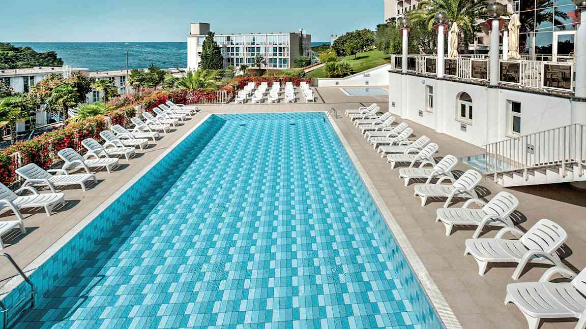 Suorakulmainen uima-allas, jonka ympärillä on aurinkotuoleja, ja taustalla meri ja rakennus. (Tekoälyn luoma teksti)