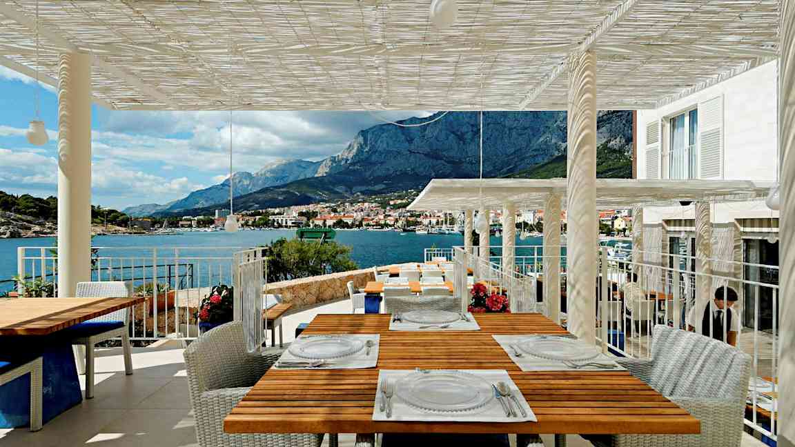 Terassi, jolta avautuu näkymä vuorille ja merelle, useat pöydät ovat katettu vieraita varten. (Tekoälyn luoma teksti)