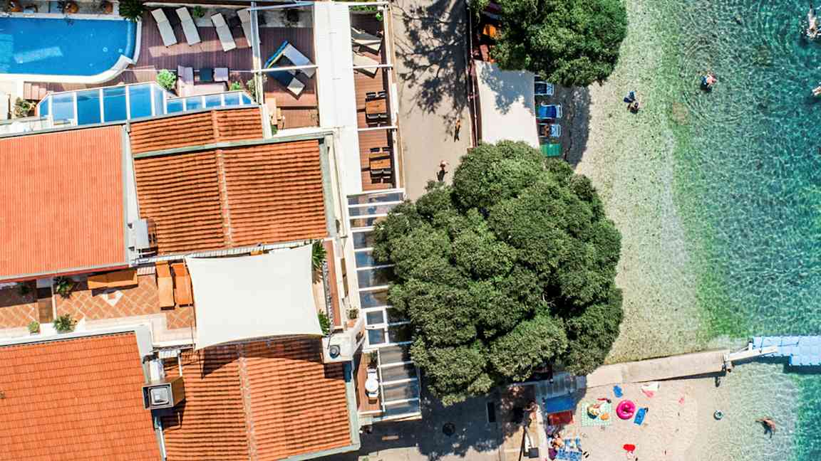 Ilmakuva rakennuksesta, jossa on uima-allas ja terassi, rannalla kirkkaan veden äärellä. (Tekoälyn luoma teksti)