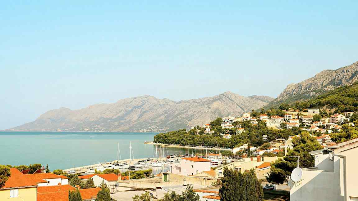 Näkymä rannikkokaupunkiin, jossa on punaisia ​​kattoja, venesatama ja vuoria taustalla. (Tekoälyn luoma teksti)