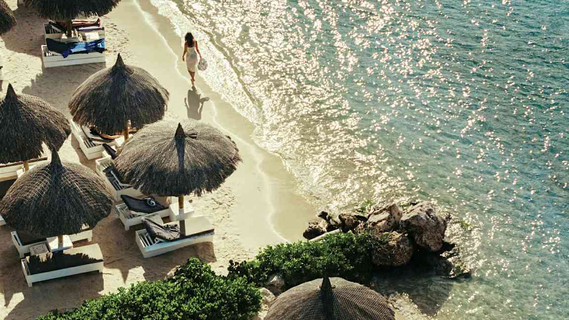 Ranta, jossa on rantavarjoja ja aurinkotuoleja, sekä henkilö kävelee veden äärellä. (Tekoälyn luoma teksti)