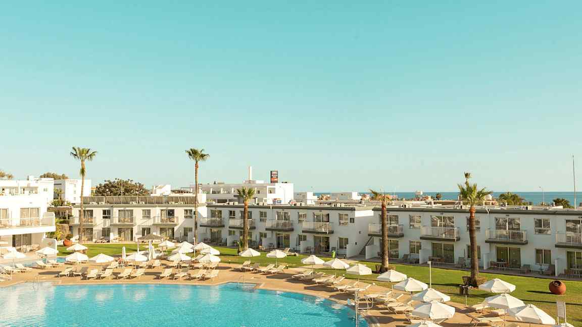 Lomakohde, jossa on uima-allas, aurinkotuoleja, varjot ja ympäröiviä rakennuksia. (Tekoälyn luoma teksti)