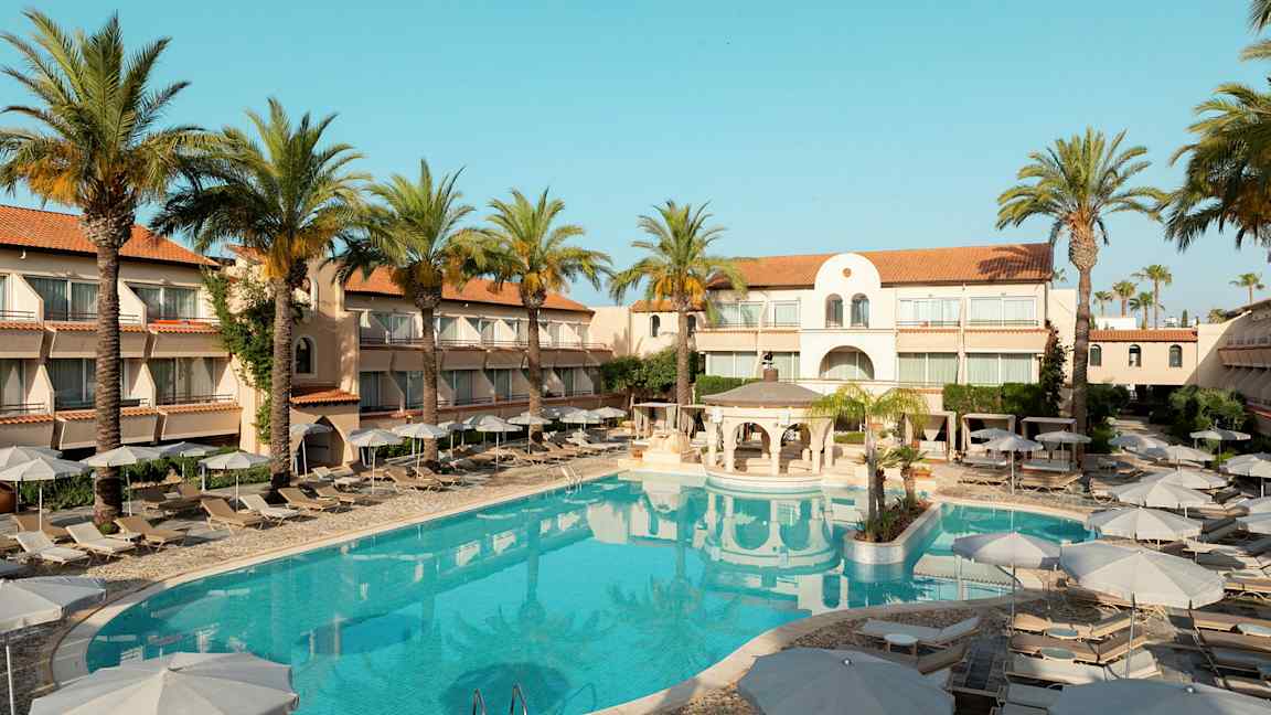 Hotellirakennus, jonka ympärillä on aurinkotuoleja ja aurinkovarjoja ulkouima-altaan äärellä, palmupuiden ympäröimänä. (Tekoälyn luoma teksti)