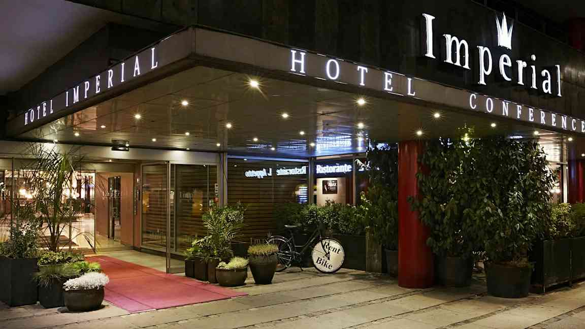 Hotel Imperial Conference -hotellin sisäänkäynti lasiovilla, ruukkukasveilla ja polkupyörällä, jossa on kyltti 'Rent a Bike'. (Tekoälyn luoma teksti)