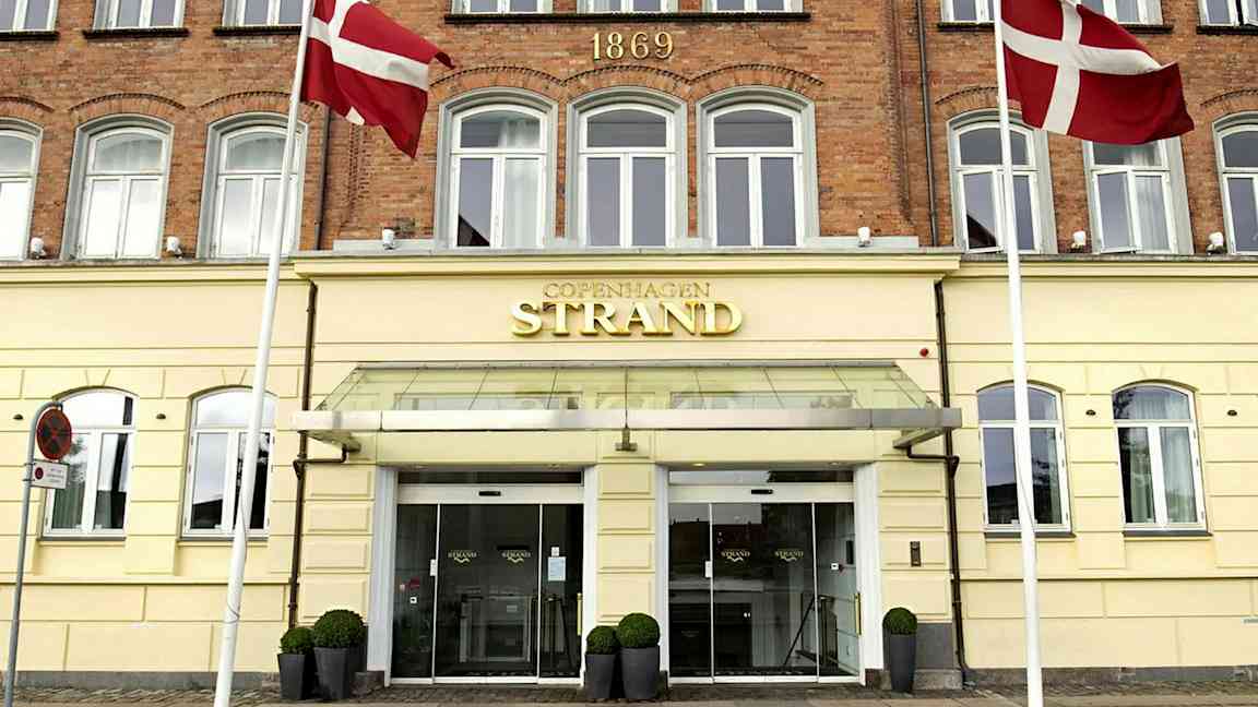 Hotellin julkisivu, jossa lukee 'Copenhagen Strand'. Tanskan lippuja näkyy sisäänkäynnin yhteydessä. (Tekoälyn luoma teksti)