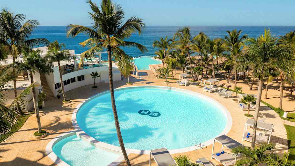 Hotellialue, jossa on suuri pyöreä uima-allas, ympäröity palmuilla, aurinkotuoleilla ja cabanoilla. Taustalla näkyy merenranta. (Tekoälyn luoma teksti)