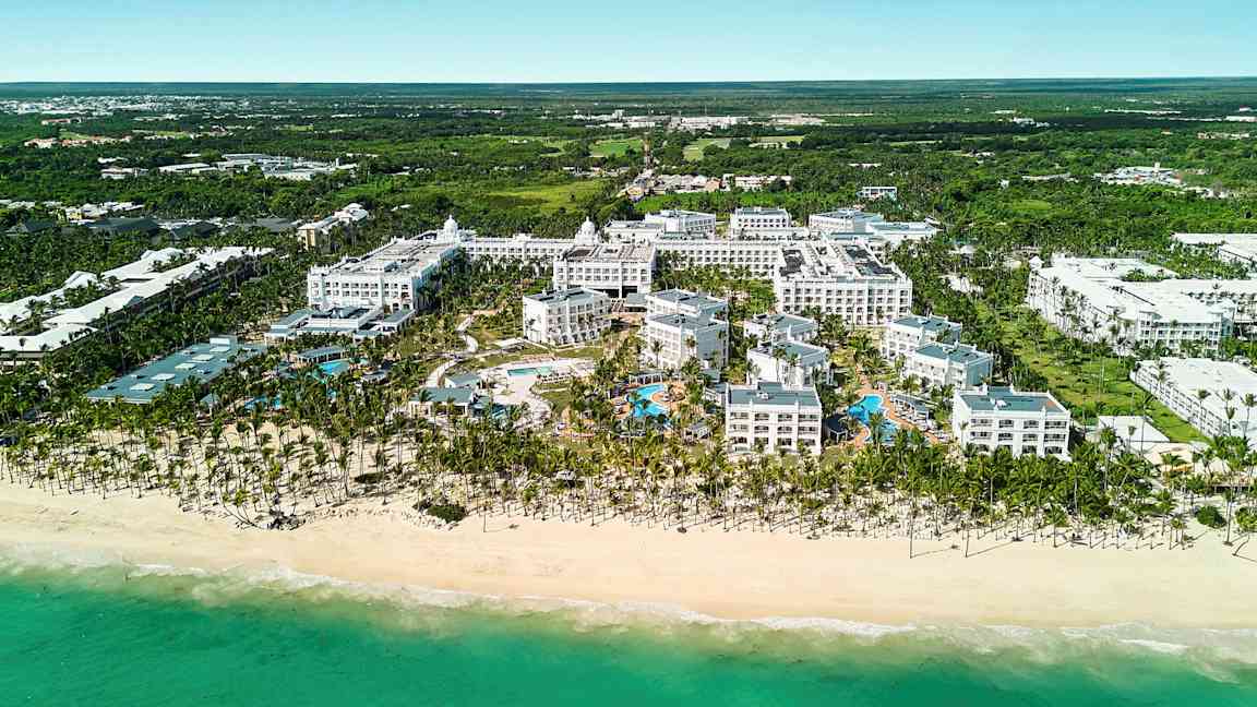 Ilmakuva hotellikompleksista rannalla sisältäen allasalueen, jota ympäröi vehreys. (Tekoälyn luoma teksti)