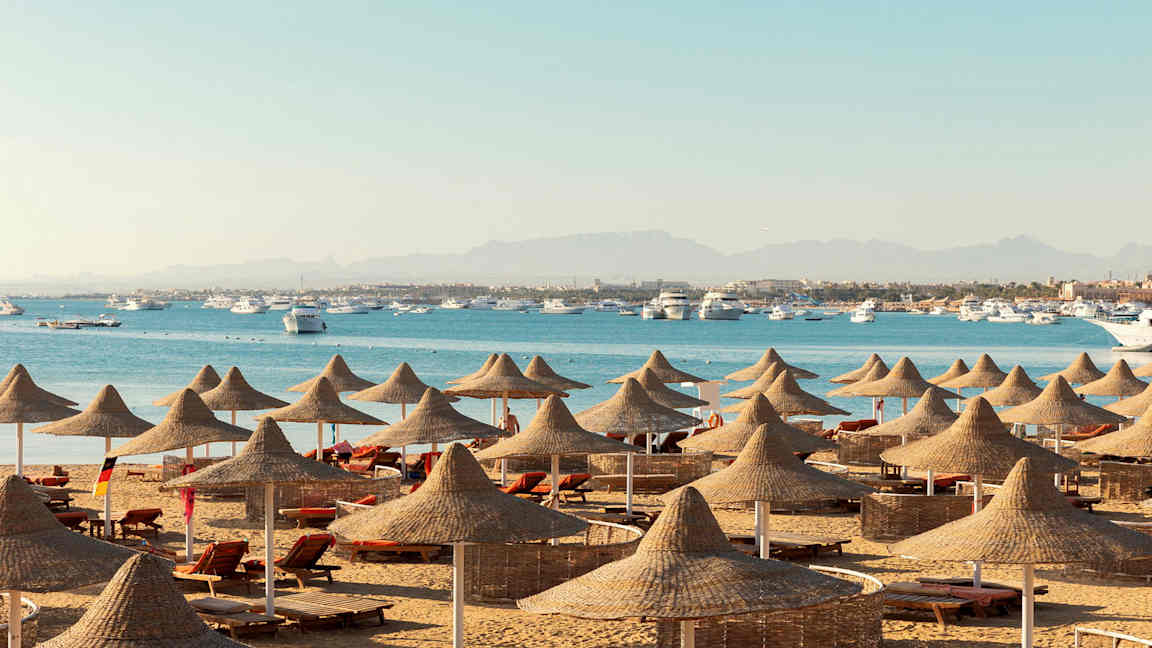 Ranta, jossa on aurinkotuoleja ja luonnonmateriaaleista valmistettuja aurinkovarjoja, taustalla meri ja veneitä. (Tekoälyn luoma teksti)