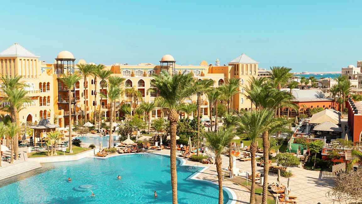 Kuva hotellialueesta, jossa on suuri uima-allas, palmuja ja lämpimän sävyisiä rakennuksia. (Tekoälyn luoma teksti)