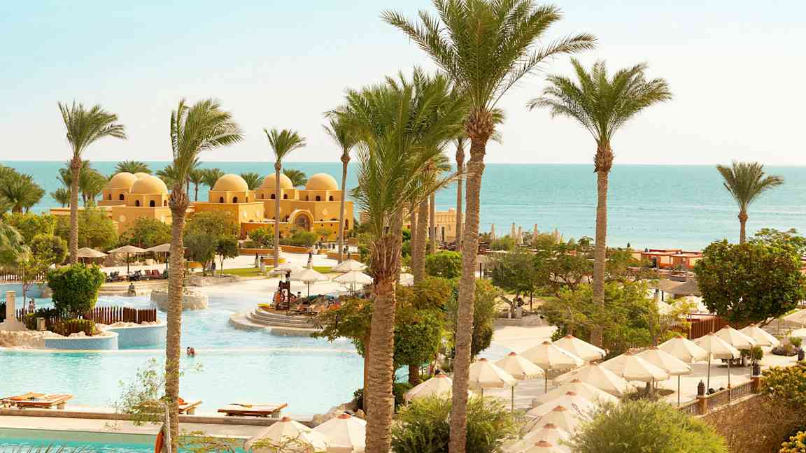 Näkymä altaalle, jossa on aurinkovarjoja, palmuja ja keltainen rakennus, jossa on kupoleja lähellä merta. (Tekoälyn luoma teksti)