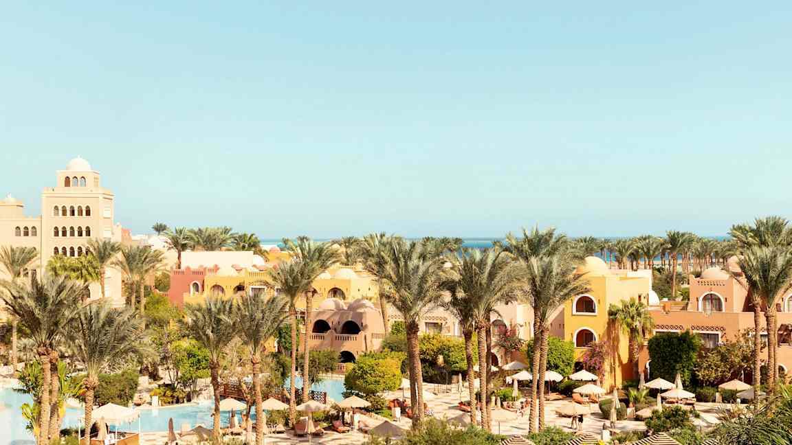 Lomakeskus, jossa on keltaisia ja oransseja rakennuksia, palmuja, vehreää kasvillisuutta sekä uima-altaita ja aurinkovarjoja. (Tekoälyn luoma teksti)