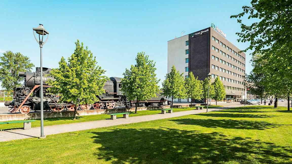 Vanha höyryveturi seisoo ulkona modernin hotellin vieressä vehreiden puiden keskellä. (Tekoälyn luoma teksti)
