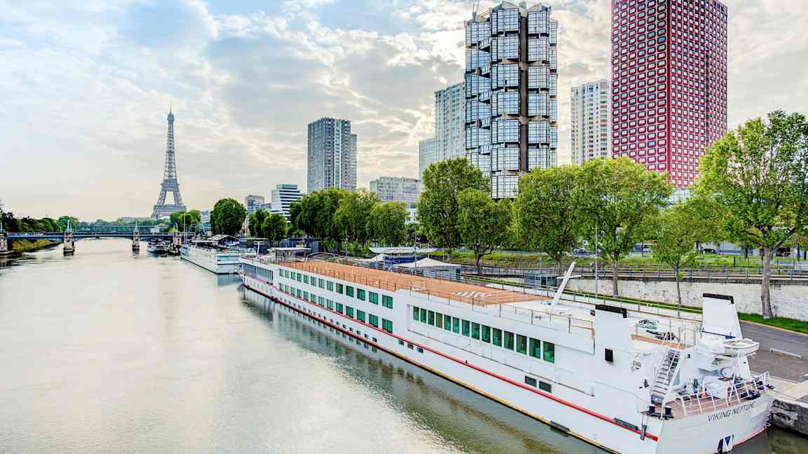 Joki, jossa vene laiturissa, moderneja rakennuksia ja Eiffel-torni taustalla. (Tekoälyn luoma teksti)