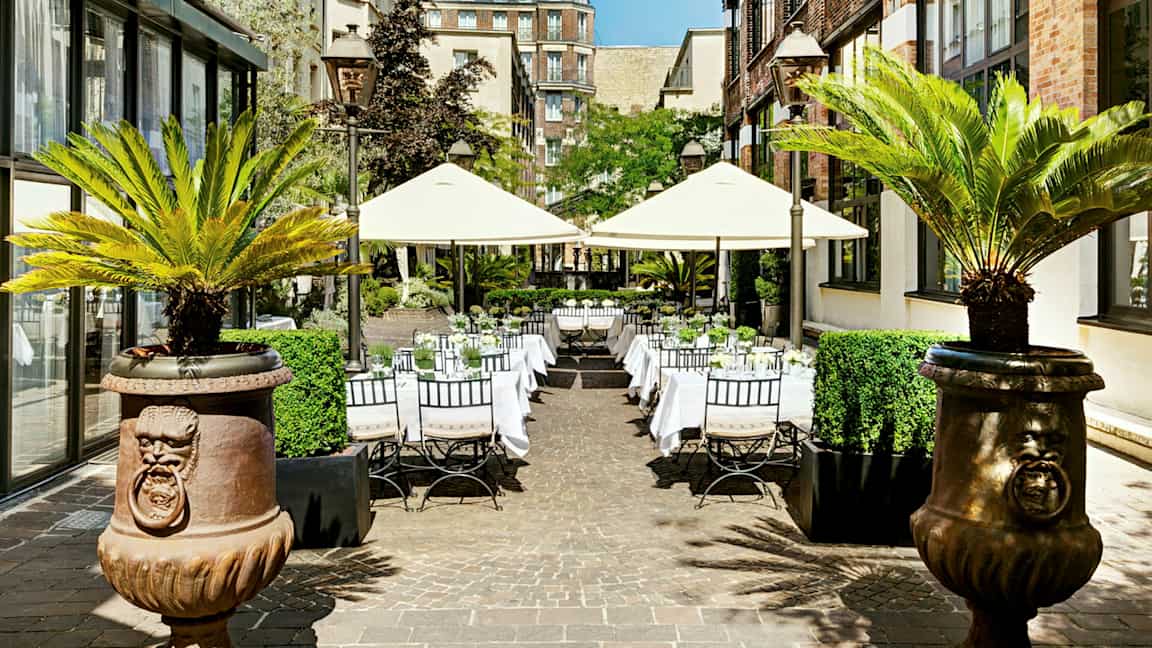 Ulkotila, jossa on pöytiä ja aurinkovarjoja, ympäröitynä rakennuksilla ja palmuilla. (Tekoälyn luoma teksti)