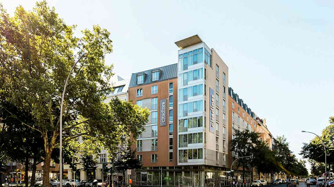 Moderni hotelli lasiseinällä, ympärillä puita ja kaupunkimaisemaa. (Tekoälyn luoma teksti)