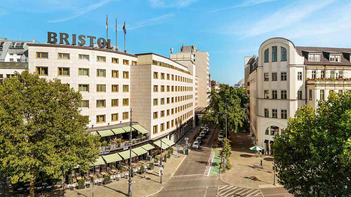 Kaupunkimaisema, jossa on rakennus, jonka katolla lukee "Bristol", ympärillä puita ja katu ulkoilmaravintoloineen. (Tekoälyn luoma teksti)