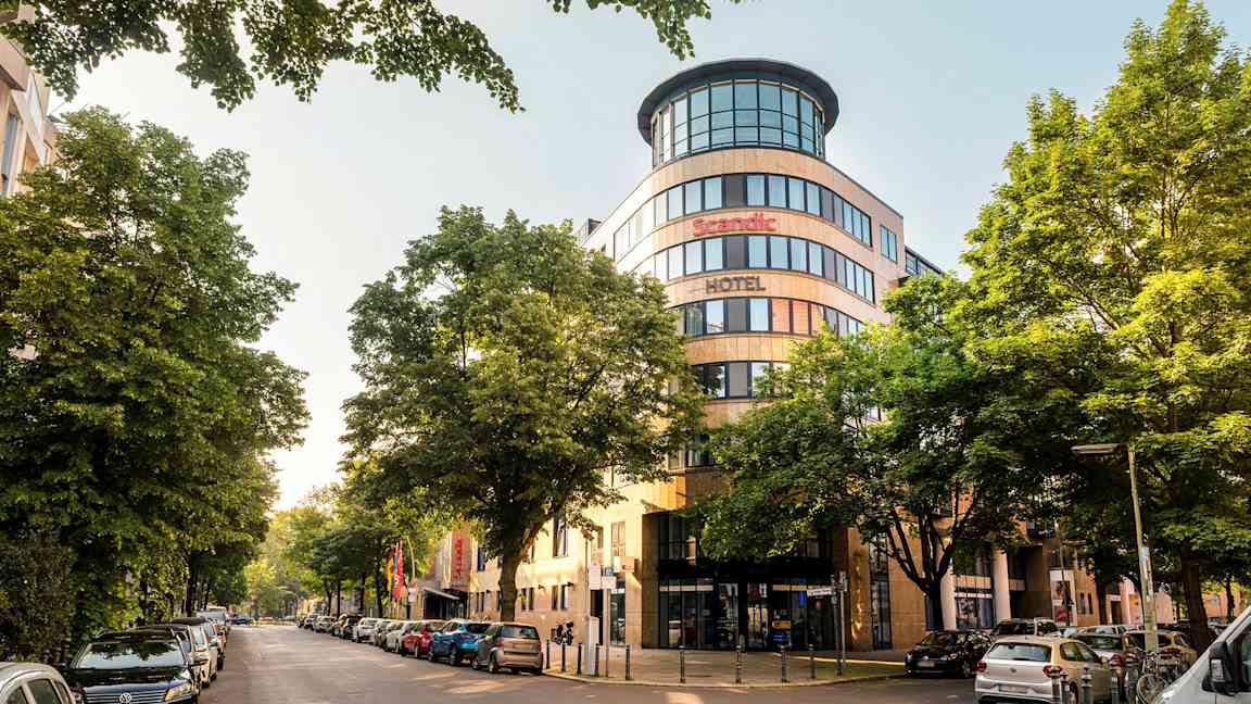 Kaupunginäkymä, jossa on Scandic-hotellin julkisivu, ympärillä puita ja pysäköityjä autoja. (Tekoälyn luoma teksti)