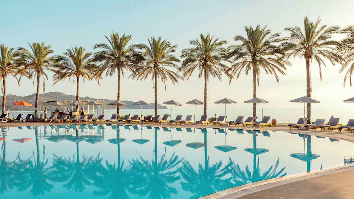 Uima-allas, jossa on aurinkotuoleja ja -varjoja, ympärillä palmuja. Meri näkyy taustalla. (Tekoälyn luoma teksti)