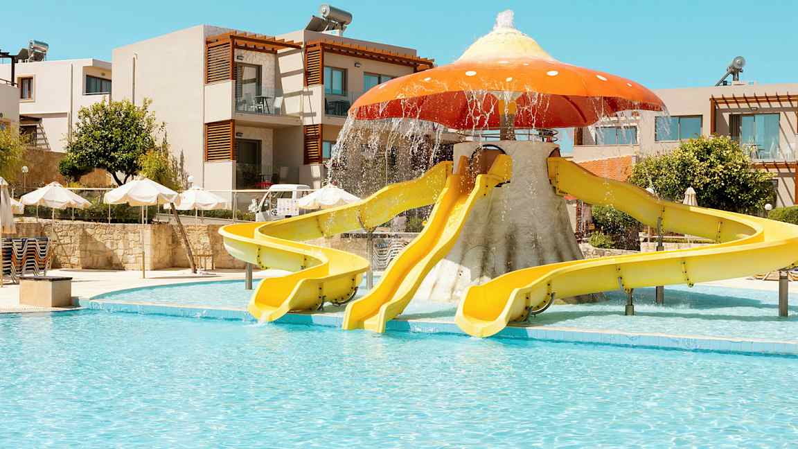 Ulkouima-altaan vieressä sienen muotoisia vesiliukumäkiä; taustalla rakennuksia ja aurinkovarjoja. (Tekoälyn luoma teksti)