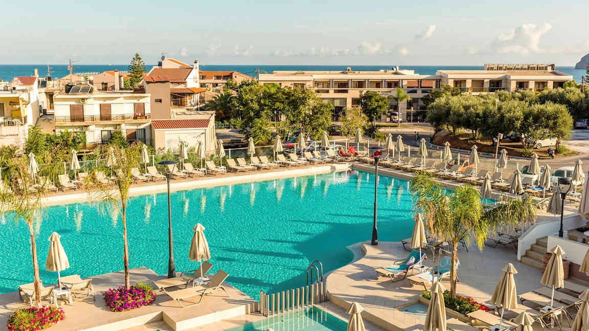 Ulkouima-allas, jota ympäröivät aurinkotuolit ja aurinkovarjot, taustalla rakennuksia ja meri. (Tekoälyn luoma teksti)