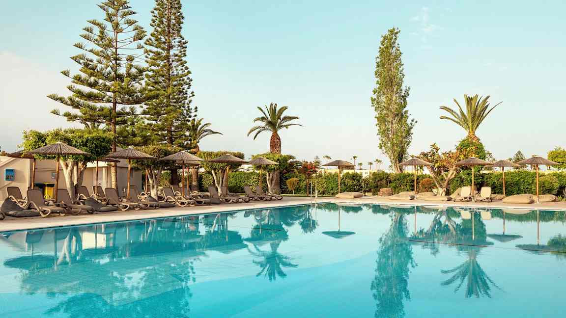 Näkymä uima-altaalle, jonka reunalla on aurinkotuoleja ja varjoja, ympärillä puita ja pensaita. (Tekoälyn luoma teksti)