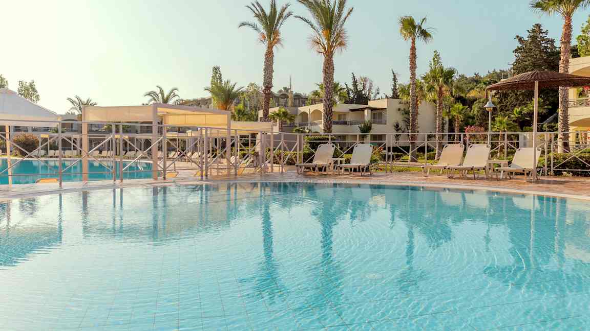 Uima-allasalue, jossa aurinkotuoleja ja aurinkovarjoja, ympäröitynä palmuilla ja rakennuksilla. (Tekoälyn luoma teksti)