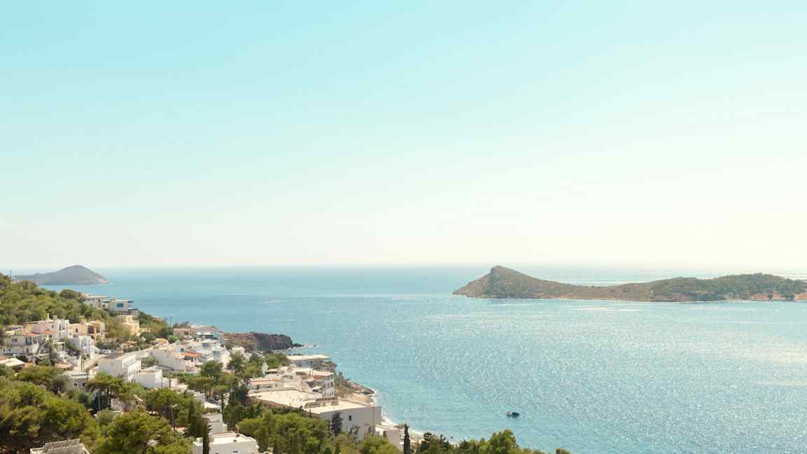 Näkymä rannikon yli, jossa valkoisia rakennuksia ja vihreitä puita, meri ja saari taustalla. (Tekoälyn luoma teksti)