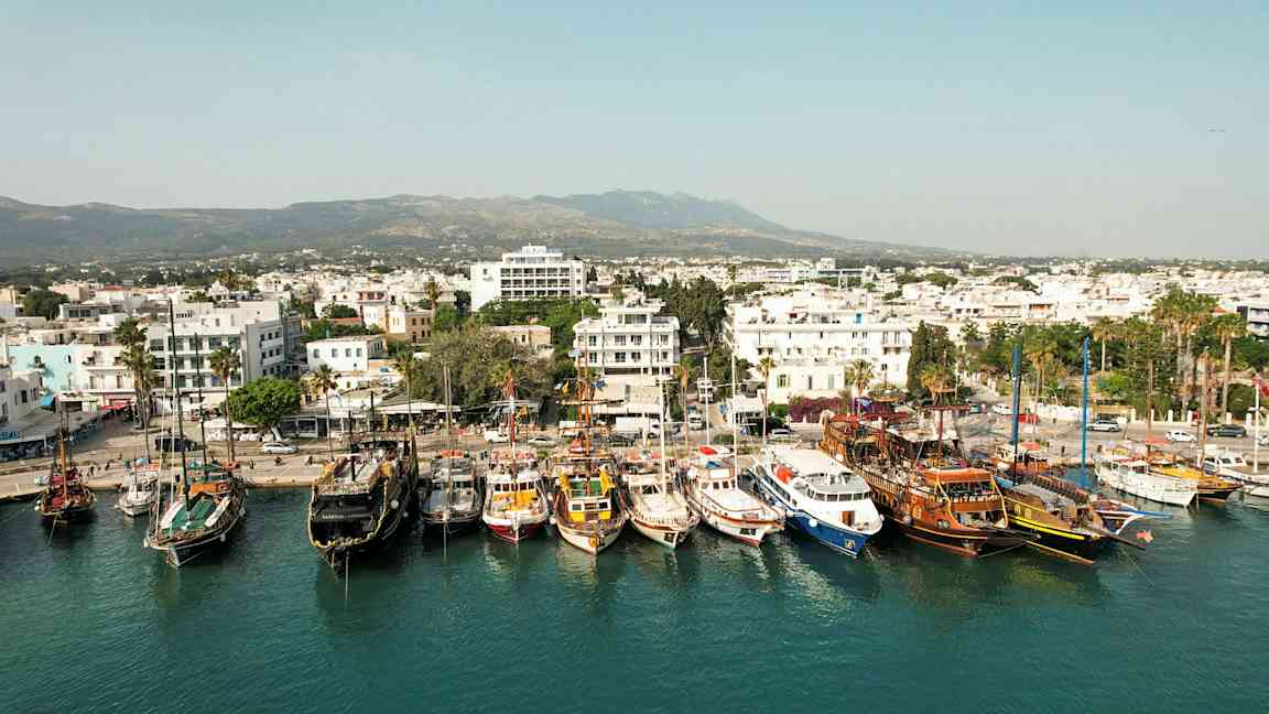 Satama, jossa useita veneitä kiinnitettynä, ja kaupunki valkoisine rakennuksineen taustalla. (Tekoälyn luoma teksti)