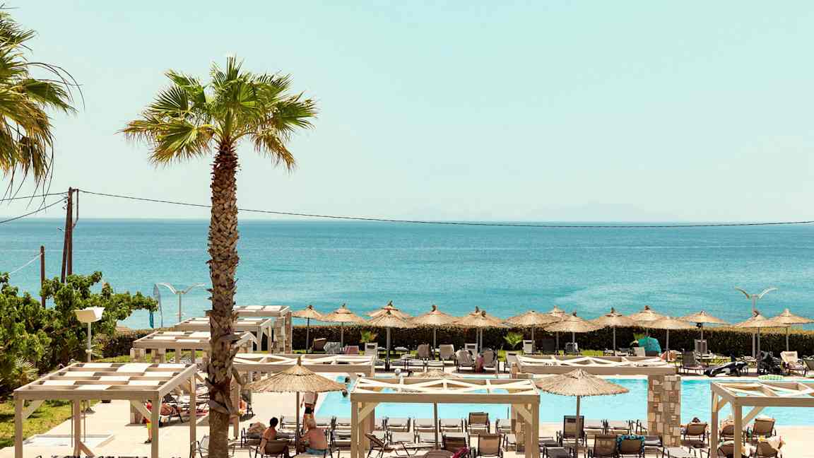 Näkymä ulkoaltaan alueelle, jossa on aurinkotuoleja, varjoja ja meri taustalla. (Tekoälyn luoma teksti)