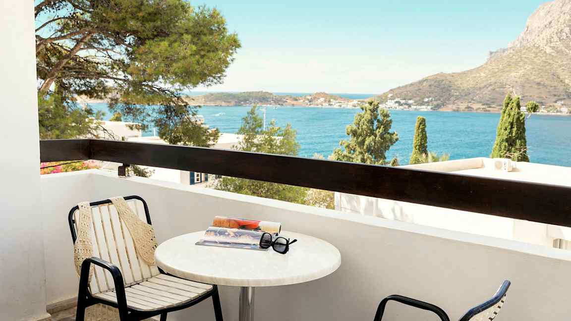 Parveke, jossa on tuoleja ja pöytä, pöydällä kirja ja aurinkolasit. Näköala merelle ja vuoristoinen tausta. (Tekoälyn luoma teksti)